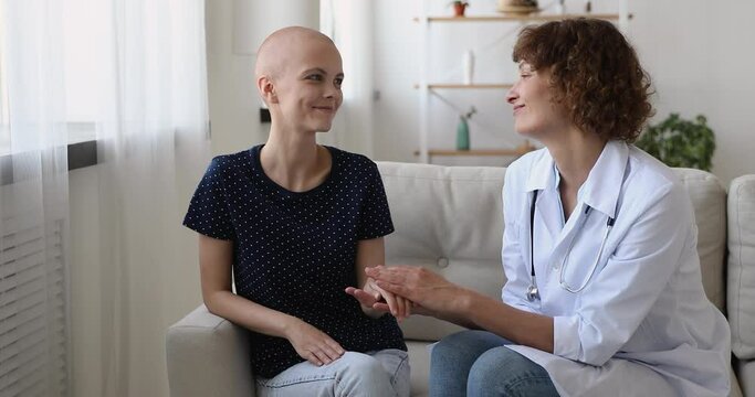 Cancer patient sit with doctor on sofa. Female oncology nurse practitioner hold strokes hand of bald ill girl express compassion showing empathy, provide psychological support and sincere care concept