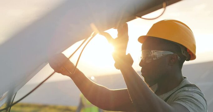 Close up of African American mechanic in protective helmet repairs solar panel with screwdriver. Technical review of modern technologies.