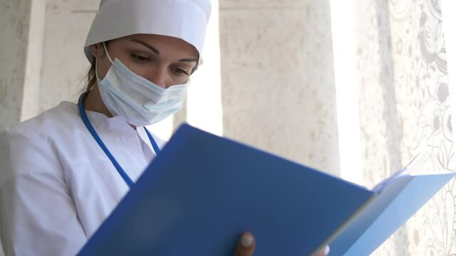 Female Doctor in medical mask standing with folder. Doctor is reading the patient's medical file