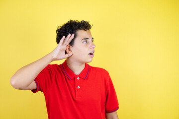 Beautiful teenager boy over isolated yellow background surprised with hand over ear listening an hearing to rumor or gossip