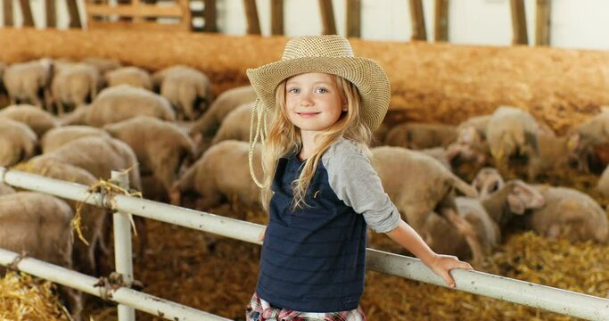 Portrait of Caucasian blonde pretty girl with fair hair standing at fence in stable and smiling to camera. Sheep flock on background. Cute little kid farmer in hat laughing in livestock barn.