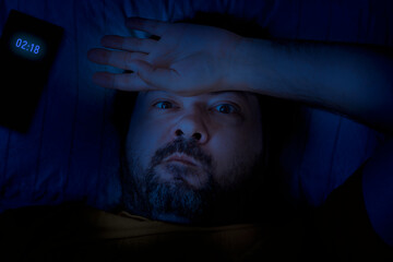 A middle-aged bearded man lies on a pillow late at night with his hand on his forehead. Concept - Insomnia and Reflections on Adult Issues