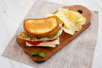 Homemade Pesto Chicken Sandwich with Potato Chips on a rustic wooden board on a white wooden background, low angle view. Close-up.