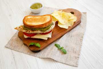 Homemade Pesto Chicken Sandwich with Potato Chips on a rustic wooden board on a white wooden background, low angle view.