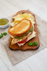 Homemade Pesto Chicken Sandwich with Potato Chips on a rustic wooden board on a white wooden background, low angle view.
