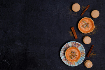 autumn orange creamy pumpkin pies for halloween with seeds, ceramic plate, cinnamon sticks, several macaroon cakes on a black background, top view, place for text, flat lay