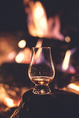Glencairn glass with a dram of scotch whisky sitting in the foreground of a blurred campfire.