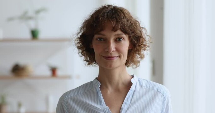 Head shot middle aged woman standing in domestic room smile looks at camera feels confident and happy. 40s pretty school teacher with curly short hairstyle, positive housewife face portrait concept