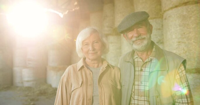 Portrait of Caucasian senior happy married couple of farmers standing together in sunlight rays, looking at each other and smiling to camera. Sunshine and hay stocks on background. Old man and woman.
