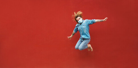 Fototapeta na wymiar Tattoo girl jumping outdoor wearing safety mask with red wall in background