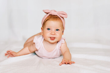 Portrait of adorable red-haired baby lying on belly, white background, wearing pink body and headband, looking straight to camera