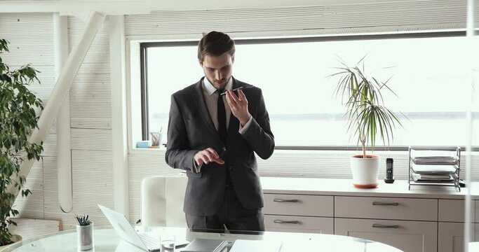 Focused young businessman using voice recognition function on smartphone, web surfing information online. Concentrated executive ceo manager in suit dictating message to client or partner in office.