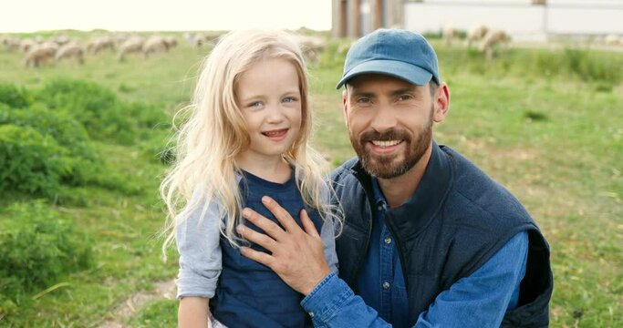 Portrait of Caucasian young handsome father standing outdoor with cute little daughter and smiling to camera in green field. Sheep flock grazing on background a pasture. Smiled man and small girl.