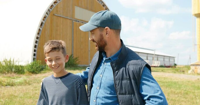 Caucasian handsome man walking outdoors with small teen boy in farm and talking, asking for advice. Male farmer speaking and discussing farming work with son. Countryside. Child and father. Discussion