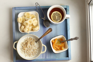 healthy breakfast on blue tray - porridge, cut banana, orange jam, lemon tea with spoon and tea strainer