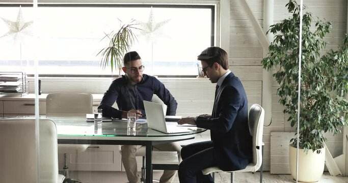 Focused young ceo leader in formal suit discussing marketing strategy with arabian male expert, sitting together at table in office. Motivated businessmen involved in working process at workplace.