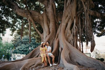 A woman sits near her boyfriend on a root of an old Valencian Ficus Macrophylla