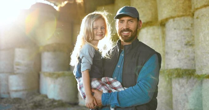 Portrait of Caucasian young handsome father holding on hands cute little daughter and smiling to camera at hay stocks in farm. Farming lifestyle. Countryside concept. Smiled man and small girl.