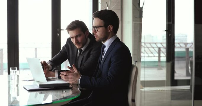 Two focused businessmen in formal wear discussing project presentation on computer, shaking hands after making agreement. Financial advisor handshaking with client after presenting profitable offer.