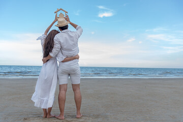 Back view of couple lover on the beach hold up a wooden home model over there head into the sky with blue sky and sea background.Make family , housing loan and real estate concept.