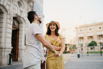 Fototapeta premium A laughing girl in a hat and a yellow dress with a plunging neckline is dancing with her boyfriend with a beard and sunglasses in the old town. A couple of tourists on the sunset in Valencia.