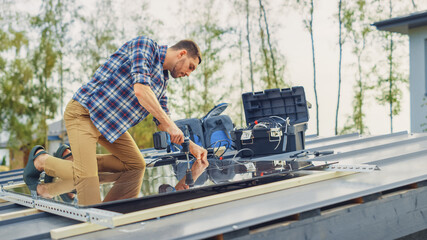 Caucasian Man in Checkered Shirt is Installing Black Reflective Solar Panels to a Metal Basis with a Drill. He Works on a House Roof on a Sunny Day. Concept of Ecological Renewable Energy at Home.