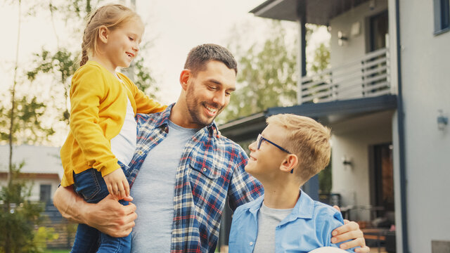 Portrait Of A Happy Family Of Three: Father, Daughter, Son. They Are Posing In Front Of Camera On A Lawn Next To Their Country House. Dad Is Holding The Girl In His Arms. Boy Is Holding A Football.