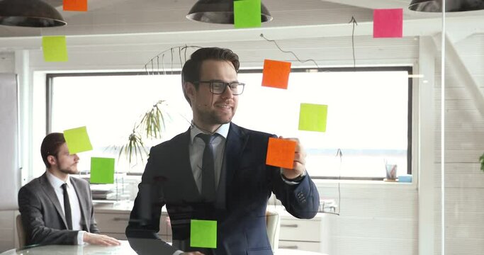 Smiling young thoughtful confident manager in formal suit and eyewear looking at glass wall, monitoring working process using agile kanban methodology. Happy boss specialist checking project workflow.