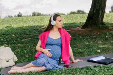 Positive dark-haired pregnant woman  listening to music in headphones with closed eyes in a park.Woman touching her belly and smiling. Prenatal care concept.Motherhood and pregnancy concept.