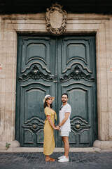 A happy girl in a hat and a dress with a plunging neckline and her boyfriend with a beard are posing near the giant doors holding each other's hand in the old town. A couple of tourists in Valencia.