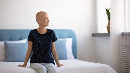 Smiling young Caucasian bald woman suffering from cancer sit on bed couch at home look in distance dreaming. Happy optimistic hairless millennial female patient with oncology think visualize recovery.