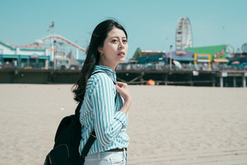 beautiful asian japanese woman backpacker walking on sandy beach outdoor on sunny day. side view of young relax charming female tourist with bag enjoy ocean with blurred amusement park in background.