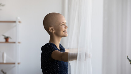 Happy young hairless sick woman suffering from oncology open curtains look in window distance dream...