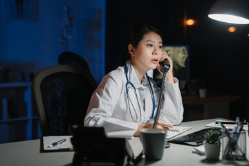 Young practitioner doctor working at 24h open clinic. asian japanese female nurse answering phone calls in late night hospital. medical staff discussing and solve problem in dark workplace midnight