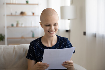 Smiling young hairless sick woman suffering from oncology sit at home read paper document with good...