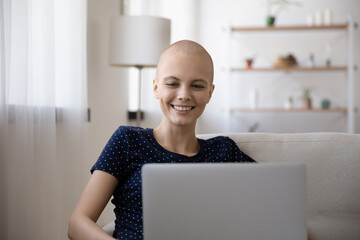 Happy young hairless sick woman struggling with oncology relax on sofa at home using modern laptop....