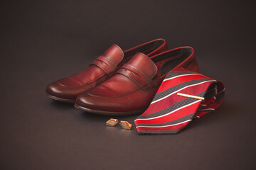 Stylish red striped rolled necktie, gold colored tie pin, cufflinks and fashionable brown men's shoes on dark background. Selective focus.