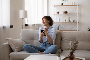 Fototapeta premium Overjoyed young woman sit on comfortable sofa in living room laugh reading good pleasant news on cellphone. Happy Caucasian female relax on couch at home have fun using smartphone gadget.