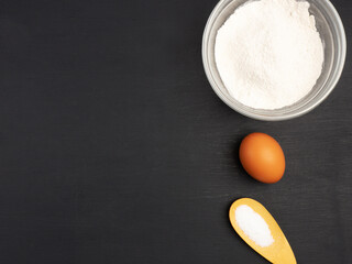 Flour, salt in spoon and egg on black wooden background