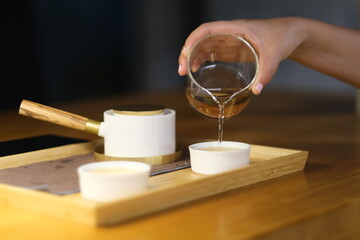 close up hand pouring tea water from traditional Chinese fair mug to tea cup. Defocused background