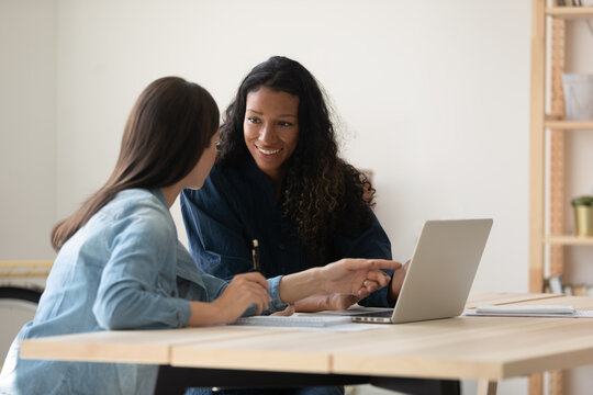 Smiling diverse colleagues working on online project together, Caucasian and African American businesswomen using laptop, discussing strategy, sharing ideas, mentor coach training new employee