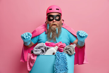 Fat bearded superhero man poses over basin full of dirty laundry wears pink cloak and helmet stares...