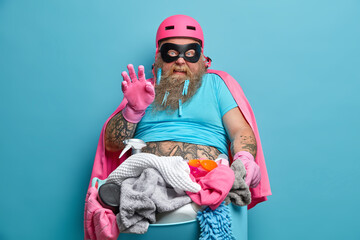 Busy superhero looks with disguise at camera holds basin of laundry and cleaning products does...