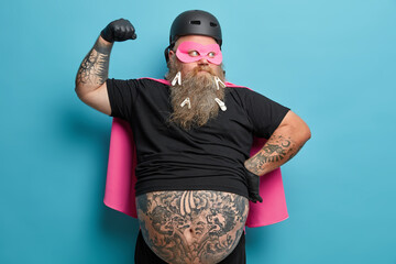 Funny bearded adult man with thick beard pretends being powerful superhero raises arm and shows...