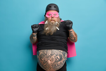 Scared fearful man has big tattooed belly wears superhero suit for costume party stares shocked at...