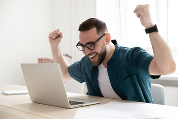 Overjoyed businessman wearing glasses looking at laptop screen, sitting at work desk in office,...