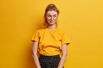 Beautiful girl with pensive mysterious expression poses against vivid yellow background dressed in casual clothes and spectacles. Young schoolgirl has pony tail concentrated aside with interest