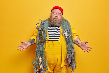 Doubtful bearded man sailor poses with fishing net spreads hands sideways feels hesitant dressed in overalls and striped shirt isolated on yellow background. Overweight fisherman goes fishing