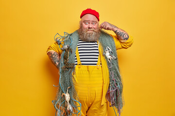 Photo of bearded sailor man curls mustache and poses indoor with fishing gears poses against yellow...