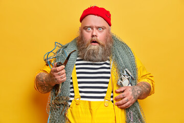 Plump bearded professional man mariner stares at camera with displeased shocked expression holds smoking pipe and goes fishing isolated on yellow background. Fatso seaman stunned to see storm at ocean - obrazy, fototapety, plakaty